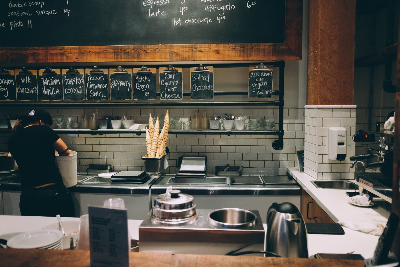 Ice cream shop interior with worker preparing cones; chalkboard menu displays various flavors above the counter.